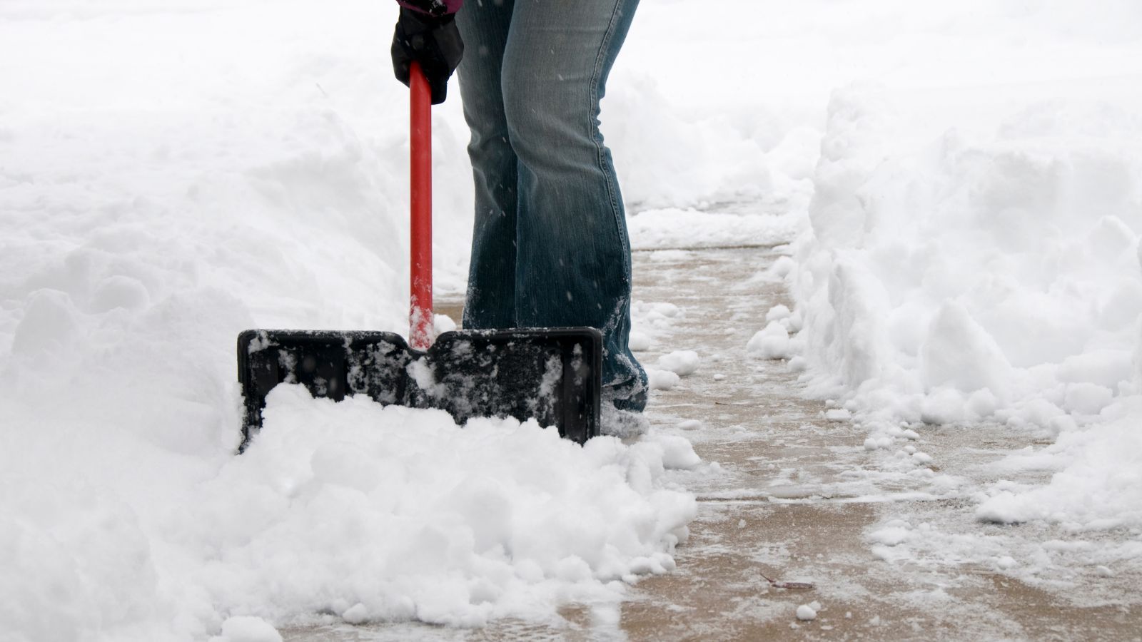 Person räumt Schnee mit einer Schneeschaufel auf einem Gehweg
