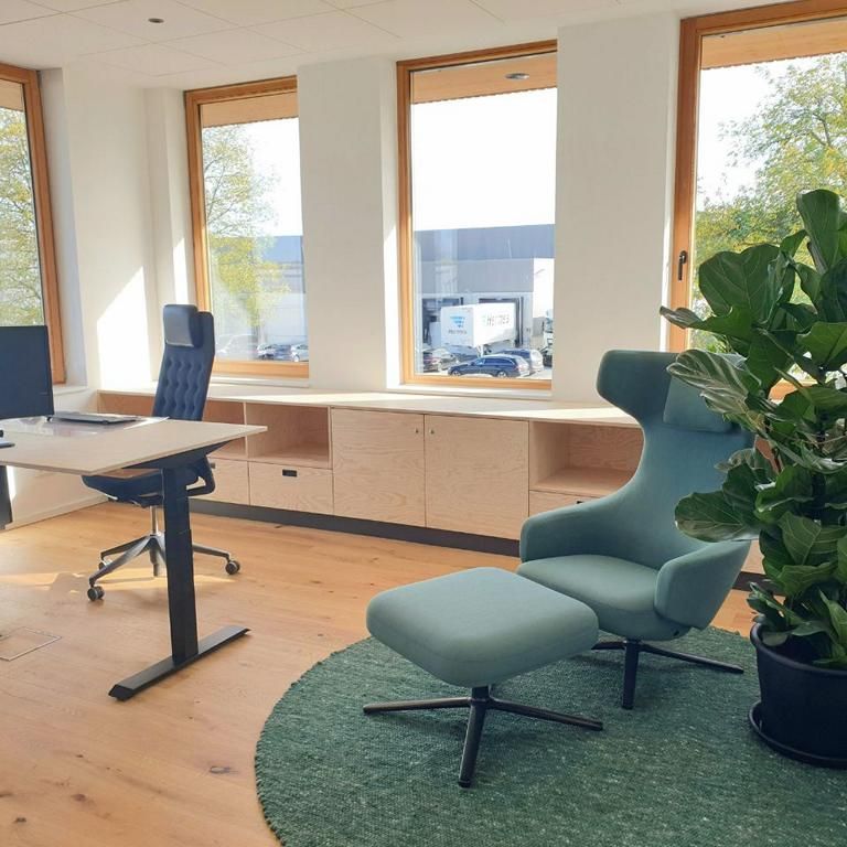Einzelbüro in einem Bürogebäude aus Holz. Man sieht Parkettboden, einen Schreibtisch aus Holz mit schwarzem Bürostuhl, Wandschränke in hellem Holz, weiße Wände und 4 große Fenster, ein grüner Sessel mit Beinablage, ein grüner Teppich und einer großen Topf-Zimmerpflanze.